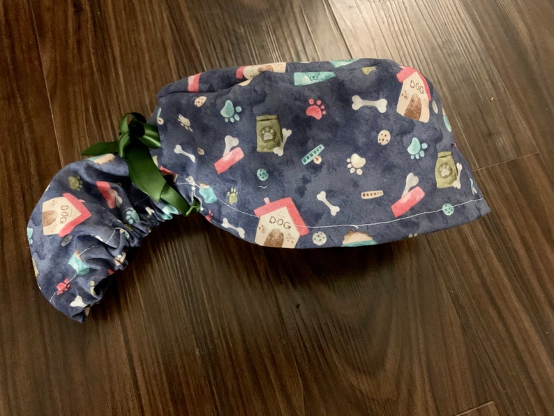 Puede incluir: Un gorro de quir&oacute;fano azul con un estampado de tem&aacute;tica canina que incluye huellas de patas, huesos, casetas para perros y bolsas de comida para perros. El gorro tiene un lazo verde.