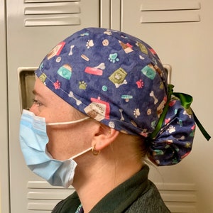 Puede incluir: Un gorro de quir&oacute;fano azul y verde con estampado y una cinta verde atada en la parte posterior. El gorro tiene varios dise&ntilde;os con tem&aacute;tica de perros, incluyendo huellas de patas, huesos y bolsas de comida para perros.