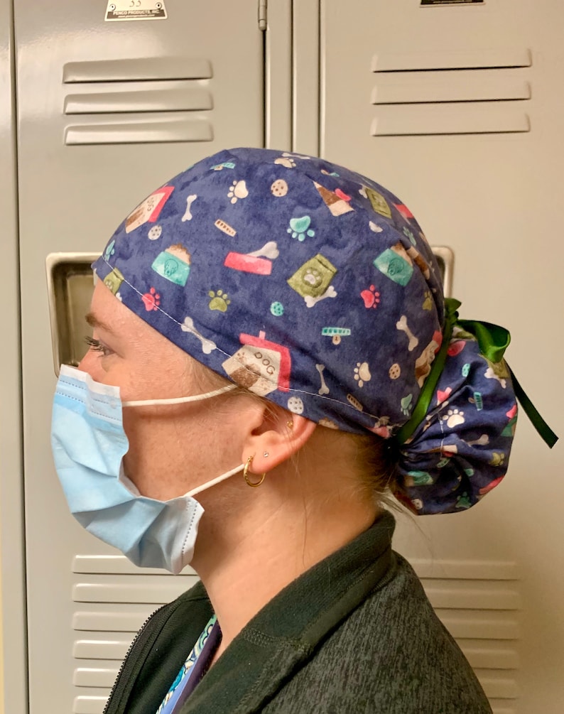 Puede incluir: Un gorro de quir&oacute;fano azul y verde con estampado y una cinta verde atada en la parte posterior. El gorro tiene varios dise&ntilde;os con tem&aacute;tica de perros, incluyendo huellas de patas, huesos y bolsas de comida para perros.