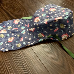 Puede incluir: Un gorro de quir&oacute;fano azul con estampado de caseta de perro y huesos. El gorro tiene una corbata verde.