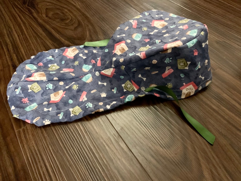 Puede incluir: Un gorro de quir&oacute;fano azul con estampado de caseta de perro y huesos. El gorro tiene una corbata verde.