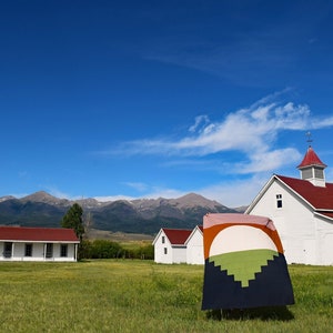 May include: A white farmhouse with a red roof and a weathervane on top, surrounded by a green field and mountains in the background. A colourful quilt is hanging in front of the farmhouse.