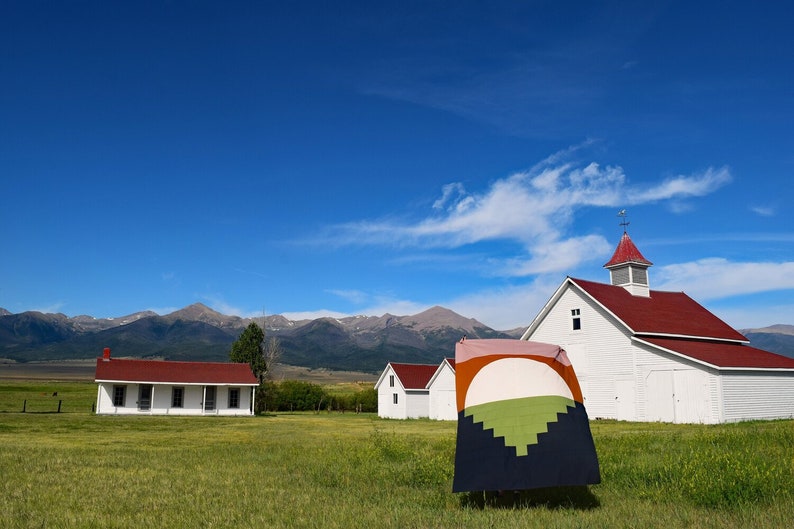May include: A white farmhouse with a red roof and a weathervane on top, surrounded by a green field and mountains in the background. A colourful quilt is hanging in front of the farmhouse.