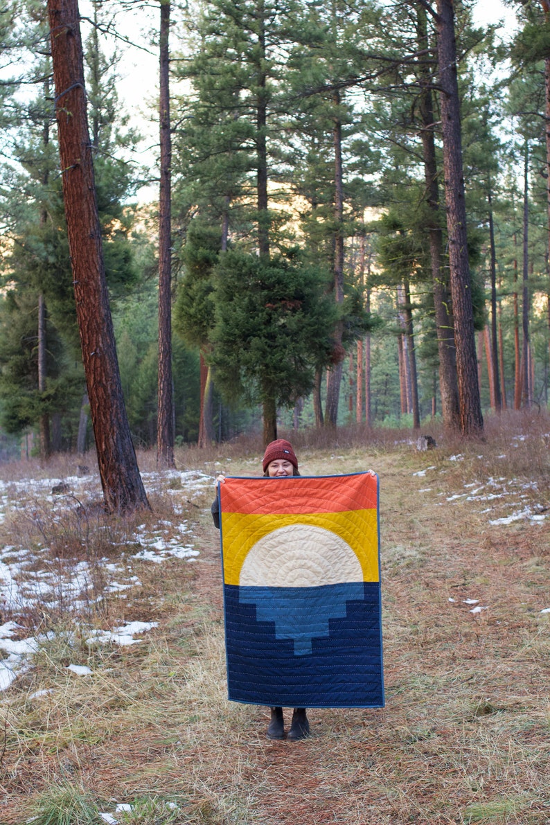 May include: A person is holding a colourful quilt with a geometric design. The quilt has a large, white circle in the centre, surrounded by orange, yellow, and blue stripes. The person is standing in a snowy forest.