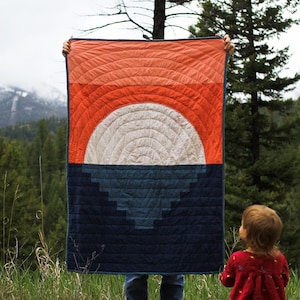 May include: A person is holding up a large quilt with a geometric design of concentric circles in shades of orange, white, and blue. The quilt is in front of a mountain range and a forest.