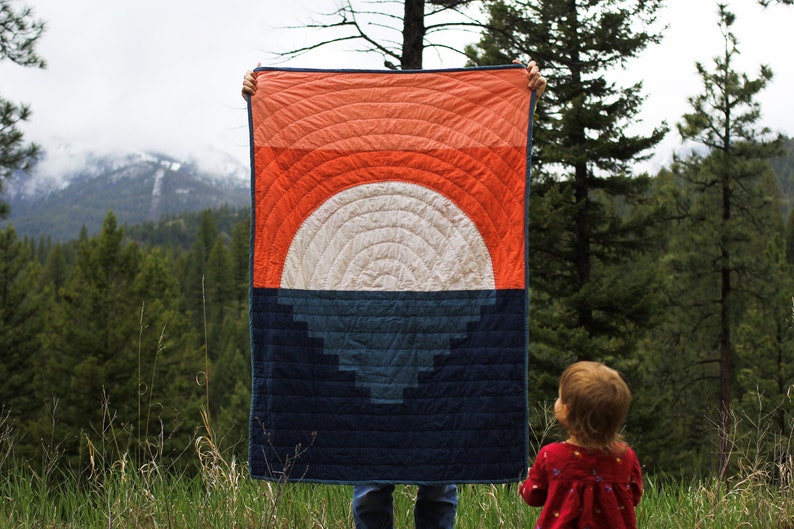 May include: A person is holding up a large quilt with a geometric design of concentric circles in shades of orange, white, and blue. The quilt is in front of a mountain range and a forest.