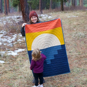 May include: A woman and a child stand in a woodland setting. The woman is holding a blue and yellow quilt with a sun design. The child is wearing a purple jumper and black trousers.