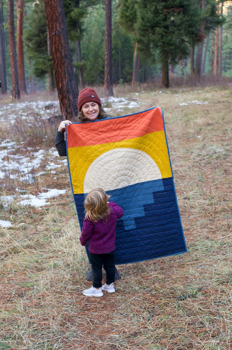 May include: A woman and a child stand in a woodland setting. The woman is holding a blue and yellow quilt with a sun design. The child is wearing a purple jumper and black trousers.