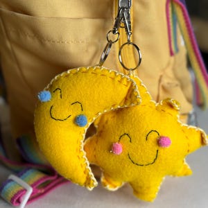 May include: A yellow felt keychain featuring a smiling sun and moon design. The sun has pink cheeks, and the moon has blue accents. The keychain is attached to a yellow backpack with a rainbow strap.