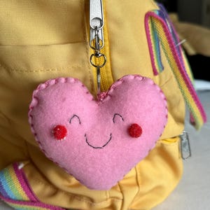May include: A pink felt heart-shaped keyring with a smiling face and red accents. The keyring is attached to a yellow backpack with a silver zip. A rainbow strap is visible.