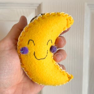 May include: A yellow felt crescent moon keyring with a smiling face and purple pom-pom cheeks. The moon is stitched with yellow thread and has a silver key ring.