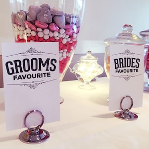 May include: Two glass jars filled with candy, one with yellow candy and the other with pink and white candy.  The jars are on a white table with two signs that say "Groom's Favorite" and "Bride's Favorite".