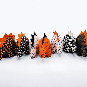 K&ouml;nnte beinhalten: Sechs Pl&uuml;sch-Dinosaurier zum Thema Halloween in verschiedenen Mustern, darunter Mais, Flederm&auml;use, Geister und Spinnweben. Die Dinosaurier sind alle in die gleiche Richtung ausgerichtet und in einer Reihe angeordnet.