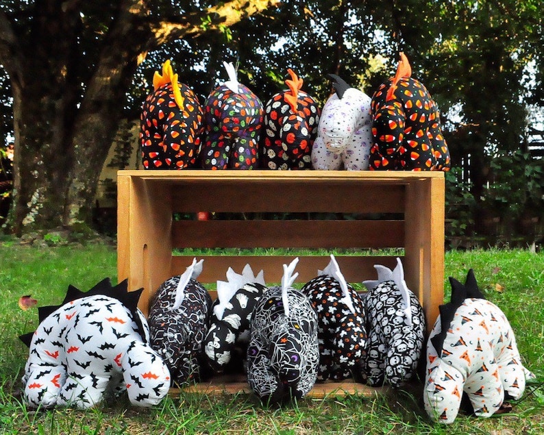 K&ouml;nnte beinhalten: Eine Sammlung von acht schwarzen und wei&szlig;en Pl&uuml;schtieren in Dinosaurierform zum Thema Halloween. Die Spielzeuge sind in einer Holzkiste angeordnet und zeigen verschiedene Muster, darunter Flederm&auml;use, Sch&auml;del und Maisbonbons.