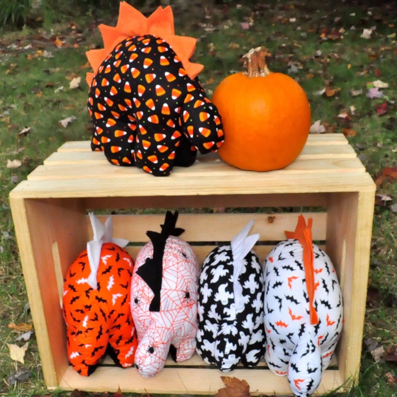 K&ouml;nnte beinhalten: F&uuml;nf Halloween-Pl&uuml;sch-Dinosaurier in einer Holzkiste. Die Dinosaurier sind mit Candy Corn, Flederm&auml;usen, Geistern und Spinnweben verziert. Ein kleiner orangefarbener K&uuml;rbis sitzt oben auf der Kiste.