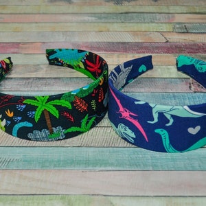 May include: Two fabric-covered headbands with dinosaur patterns. One headband is black with green, blue, and red dinosaurs and palm trees. The other headband is blue with light blue, green, and pink dinosaurs.