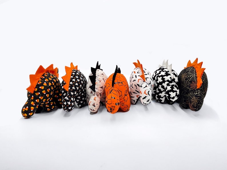 K&ouml;nnte beinhalten: Sechs Pl&uuml;sch-Dinosaurier zum Thema Halloween in verschiedenen Mustern, darunter Mais, Flederm&auml;use, Geister und Spinnweben. Die Dinosaurier sind alle in die gleiche Richtung ausgerichtet und in einer Reihe angeordnet.