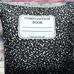 Peut inclure: Un coussin carré avec un motif de couverture de cahier noir et blanc. Le coussin présente le texte "COMPOSITION BOOK" dans un rectangle blanc, avec des lignes en dessous et le texte "wide ruled • 100 pages". Le coussin a un motif tacheté.