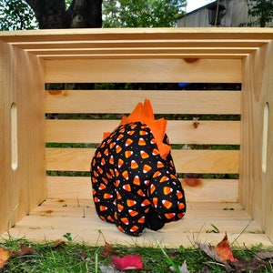 K&ouml;nnte beinhalten: Ein schwarzes und oranges Stoff-Dinosaurierspielzeug mit Candy-Corn-Druck und orangefarbenen Stacheln. Das Spielzeug sitzt in einer Holzkiste.