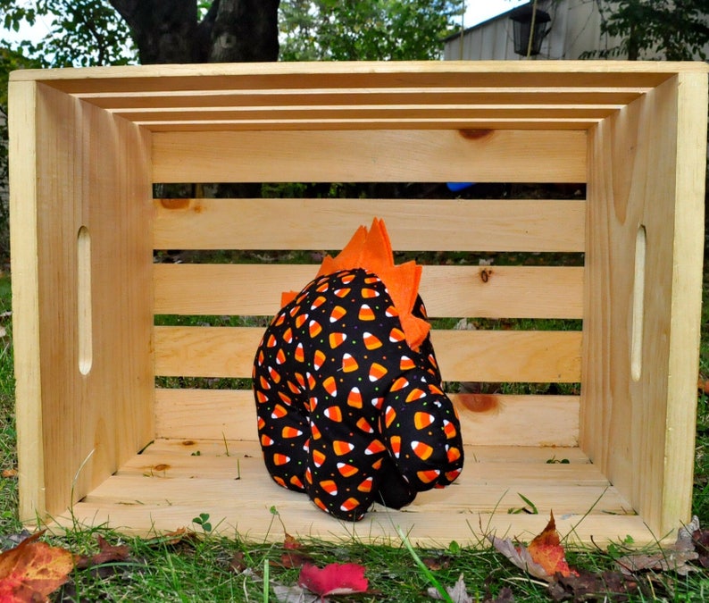 K&ouml;nnte beinhalten: Ein schwarzes und oranges Stoff-Dinosaurierspielzeug mit Candy-Corn-Druck und orangefarbenen Stacheln. Das Spielzeug sitzt in einer Holzkiste.