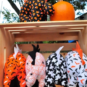 K&ouml;nnte beinhalten: Vier Halloween-Pl&uuml;schtiere in einer Holzkiste. Die Spielzeuge sind in Form eines Geistes, einer Spinne, einer Fledermaus und eines Mais-S&uuml;&szlig;igkeitens. Die Spielzeuge sind alle schwarz, wei&szlig; und orange.