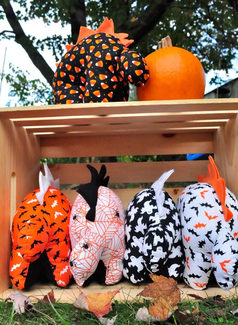 K&ouml;nnte beinhalten: Vier Halloween-Pl&uuml;schtiere in einer Holzkiste. Die Spielzeuge sind in Form eines Geistes, einer Spinne, einer Fledermaus und eines Mais-S&uuml;&szlig;igkeitens. Die Spielzeuge sind alle schwarz, wei&szlig; und orange.