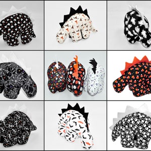 K&ouml;nnte beinhalten: Neun Pl&uuml;sch-Dinosaurier-Spielzeuge mit verschiedenen Halloween-Motiven. Die Dinosaurier sind schwarz und wei&szlig; mit Mustern von Geistern, Flederm&auml;usen, Zuckermais, Sch&auml;deln und Spinnweben.