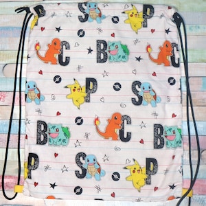 Op de afbeelding: Een witte trekkoordtas met een herhalend patroon van Pokémon-personages, waaronder Pikachu, Bulbasaur en Charmander. De tas heeft de letters "B", "P", "S" en "C" in een zwart-witte grunge-lettertype.