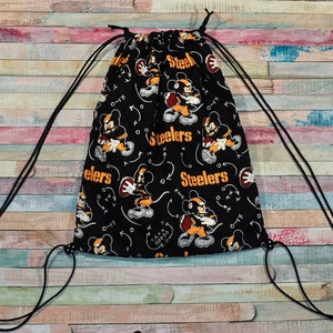May include: Black drawstring bag with a black and yellow pattern featuring Mickey Mouse playing football. The bag has the word "Steelers" repeated on the fabric.