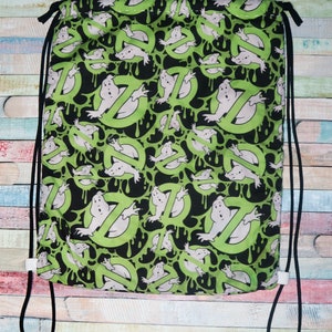 May include: A drawstring backpack featuring a repeating pattern of the Ghostbusters logo and ghost figures on a black and green background. The bag has black drawstrings and white plastic cord locks. The bag is made of fabric.