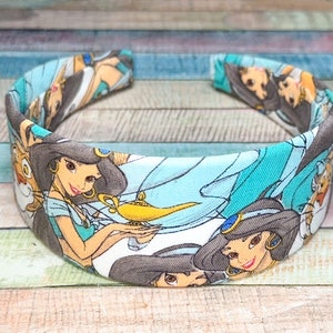 May include: A blue headband with a white background featuring a repeating pattern of Jasmine from the Disney movie Aladdin. The pattern includes Jasmine, Abu, the Genie's lamp, and other characters from the movie.