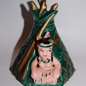 May include: A ceramic figurine depicting a Native American woman seated in front of a green and gold teepee. The figurine is painted in shades of pink, green, and gold, with intricate details on the teepee and the woman's attire.