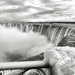 Op de afbeelding: Zwart-wit foto van de Niagara Falls, met het waterval en de mist. De afbeelding toont de kracht van de watervallen, met een gedeeltelijk bevroren reling op de voorgrond, die textuur en detail aan de scène toevoegt.