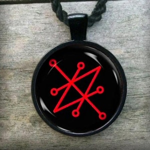 Op de afbeelding: Een zwarte ketting met een rode zegel op een zwarte achtergrond. De zegel is een geometrisch ontwerp met vijf punten en een cirkel op elk punt.