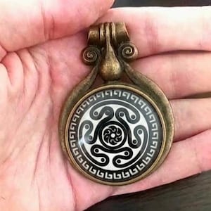 May include: A circular pendant with an antique bronze-toned metal frame featuring a decorative bail. The center inlay is white with a black spiral triskelion design surrounded by a black and white geometric Greek key border pattern.
