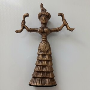 May include: Bronze-coloured figurine of a woman with outstretched arms, holding curved objects. She wears a tiered skirt and a headdress. The statue is likely a decorative item or a collectible.