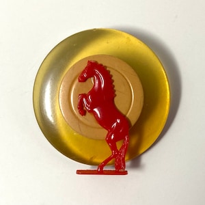 May include: A circular brooch featuring a translucent yellow outer disc and a solid beige inner disc. A bright red plastic horse figure in a rearing position is attached to the front, standing on a small rectangular base.