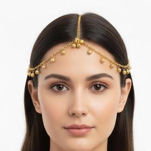 Könnte beinhalten: Ein goldfarbener Kopfschmuck mit einer zarten Kette, die auf der Stirn aufliegt und mit kleinen, gleichmäßig verteilten kugelförmigen Perlen verziert ist. Das Accessoire wird zu einem hellrosa Kleidungsstück mit goldener Stickerei getragen.