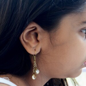 May include: A young girl with long dark hair wearing a pair of gold dangle earrings with clear crystal stones.