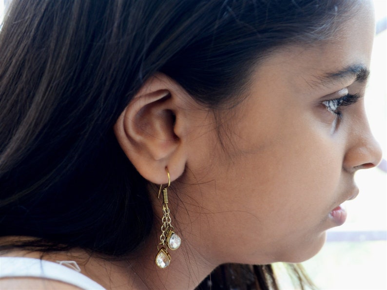 May include: A young girl with long dark hair wearing a pair of gold dangle earrings with clear crystal stones.