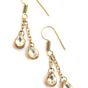 May include: Gold tone dangle earrings with clear teardrop shaped crystals. The earrings have two chains with a crystal at the end of each chain.