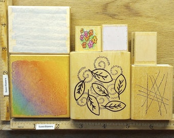 May include: A collection of wooden stamps with various designs. The stamps include a watercolor wash, floral pattern, leaf design, and a series of intersecting lines. The stamps are arranged on a wooden surface with a ruler for scale. The text "SaverStamps" is visible.