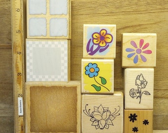 May include: A collection of wooden stamps with various designs, including flowers, a checkered pattern, and a blank square. The stamps are arranged on a wooden surface next to a ruler marked in inches. The stamps are in various colors.