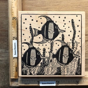 May include: A wooden rubber stamp with a black and white design of three fish swimming in a coral reef. The stamp is approximately 2 inches square.