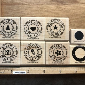 May include: Eight wooden rubber stamps with different designs and sayings. The stamps are in a grid of two rows and four columns. The sayings on the stamps include "Happy Holidays", "For You With Love", "Wishing You The Best", "Enjoy Your Day", "Welcome Little One", "Thanks So Much", and two blank stamps with a circle and a star.