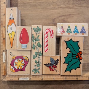 May include: A collection of eight wooden rubber stamps featuring Christmas designs including ornaments, a light bulb, mistletoe, a candy cane, four small trees, and holly leaves, arranged against a ruler showing 6 inches.