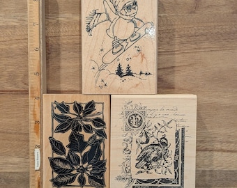 May include: Three wooden stamps with black ink designs. One stamp features a snowman snowboarding, another a poinsettia, and the third a bird with text. The stamps are on a wooden surface.