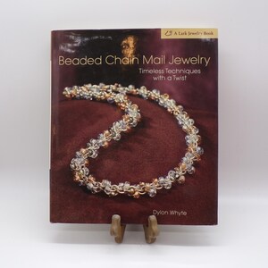 May include: A book titled "Beaded Chain Mail Jewelry" with a necklace on the cover. The cover is a deep burgundy color with the title in white and the author's name, Dylon Whyte, at the bottom. The book is propped up on a wooden stand.