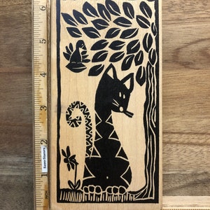 May include: A wooden rubber stamp with a black and white design of a cat sitting under a tree with leaves. A bird is perched on a branch of the tree. The stamp measures approximately 2 inches by 3 inches.