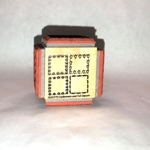 May include: A wooden stamp with two different designs. The top design is a square frame with a border of small hearts. The bottom design is a square frame with a border of small circles. The stamp is labeled "002779 Halloween and Fall Square".